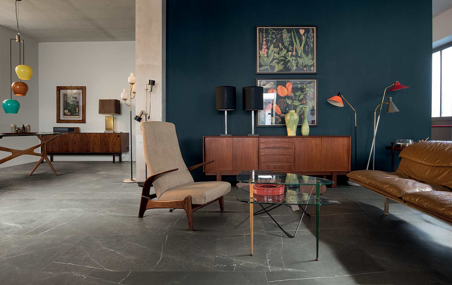 Rustykalny salon z podłogą wyłożoną brązowymi płytkami imitującymi kamień Amani Bronze z jedną ścianą niebieską, dwoma drewnianymi szafkami, stołem i szklanym stolikiem, fotelem, kanapą, lampami wiszącymi i stojącymi, obrazami na ścianie i ozdobami