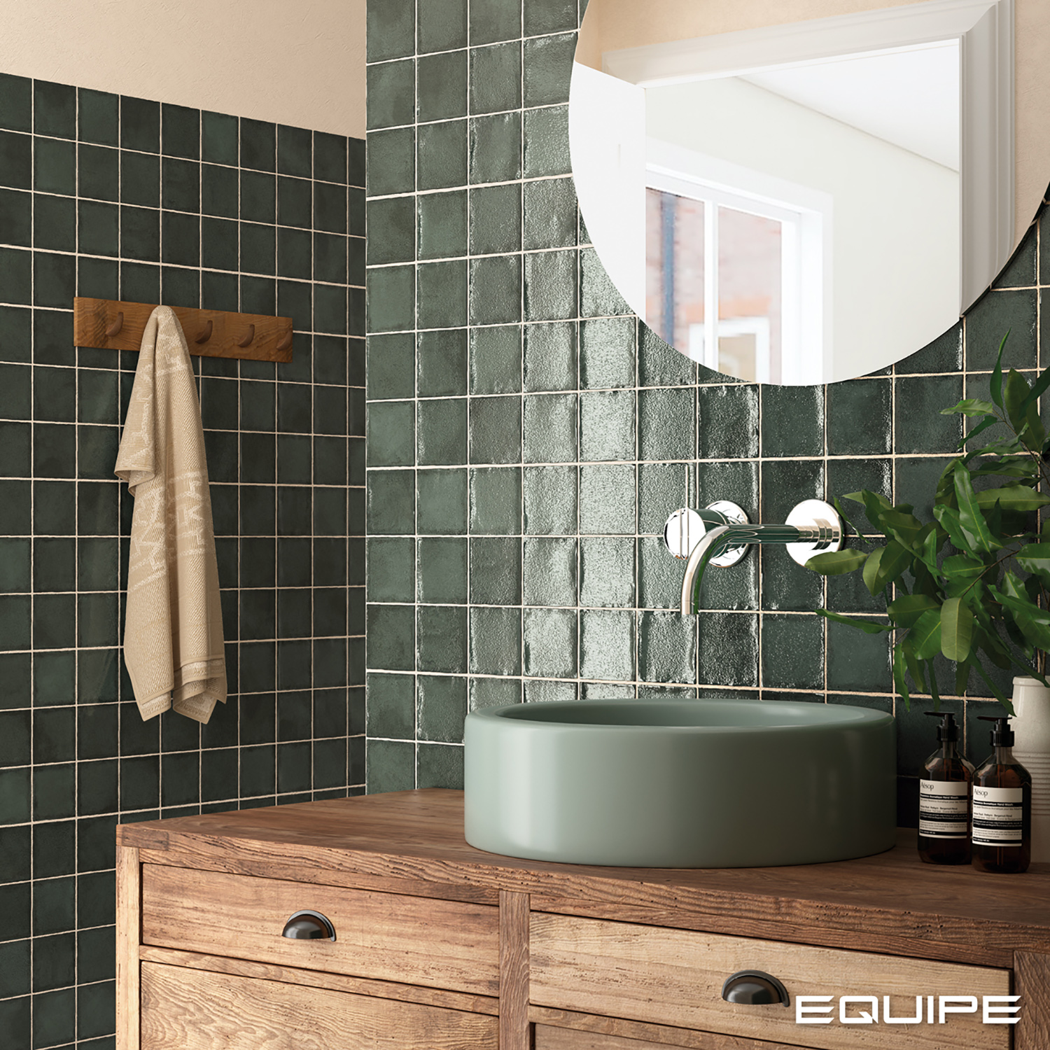 Łazienka ze ścianą wyłożoną zielonymi cegiełkami w formie kwadratu, z drewnianą szafką, zieloną umywalką nablatową, okrągłym lustrem, kwiatem i drewnianym wieszakiem z ręcznikiem