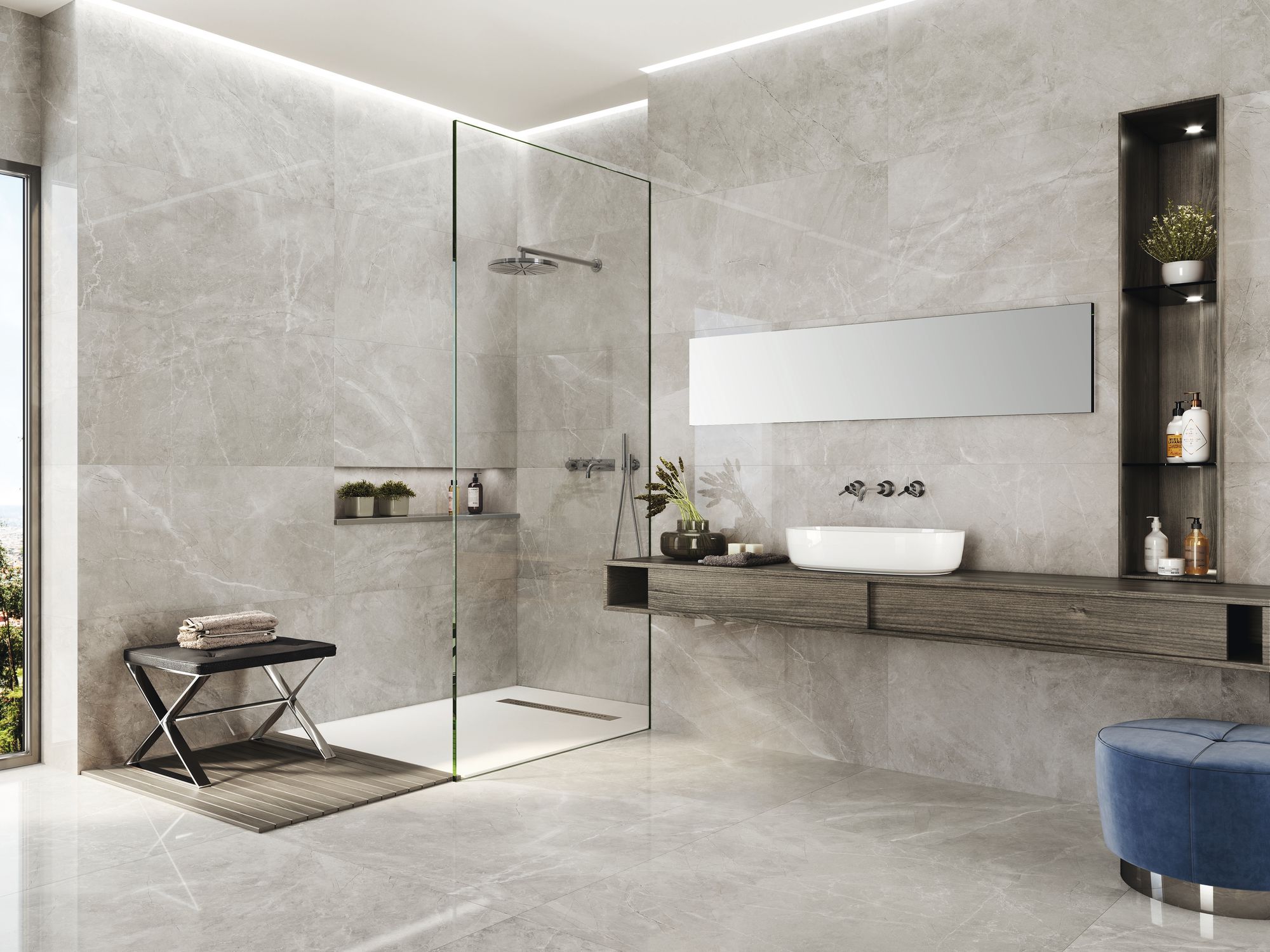 Szara łazienka wyłożona płytkami imitującymi kamień z kabiną prysznicową, wiszącą półką drewnianą z umywalką nablatową, prostokątnym wąskim lustrem, półką w ścianie z kosmetykami, niebieską pufą i niskim siedziskiem