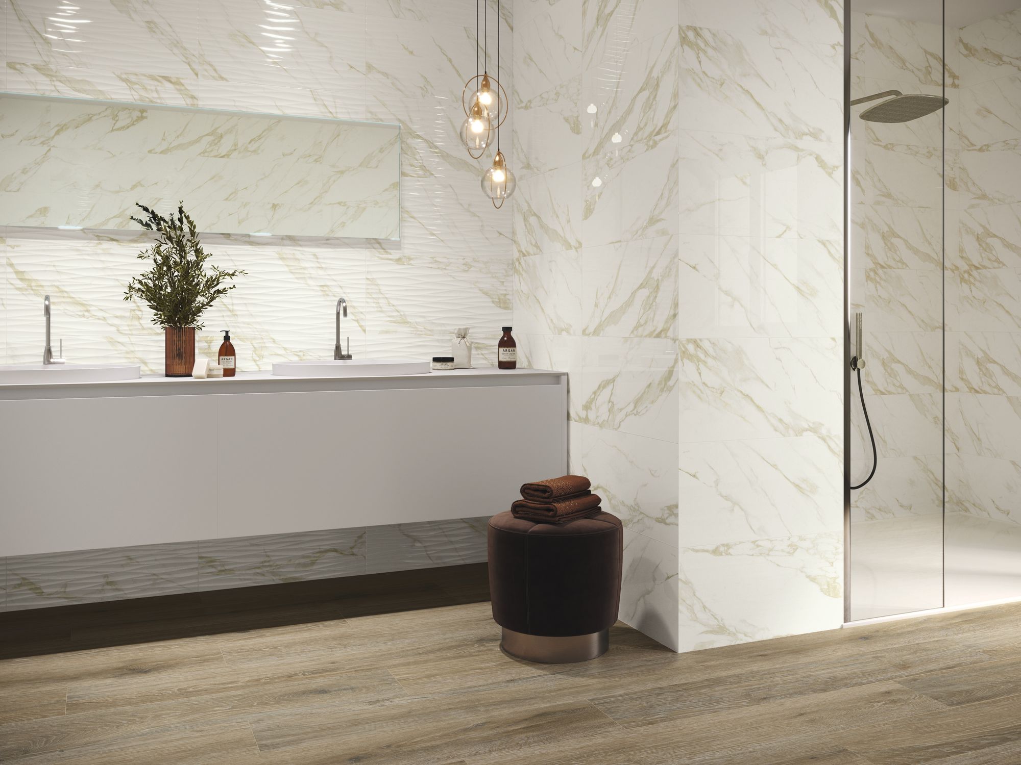 Jasna łazienka ze ścianą wyłożoną płytkami imitującymi marmur i drewnopodobną podłogą, z kabiną prysznicową, długą, białą szafką wiszącą z dwiema umywalkami, długim, poziomym lustrem, lampą wiszącą i brązową pufą