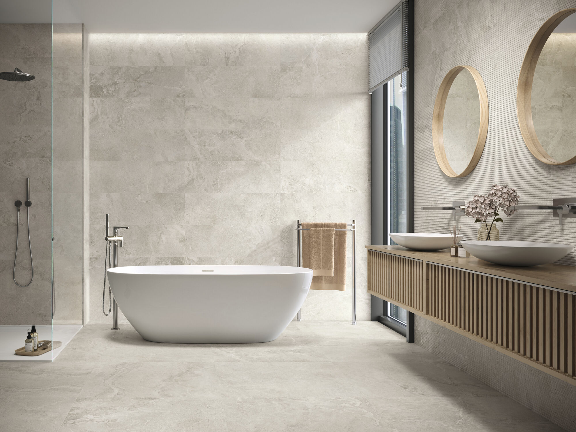 Łazienka wyłożona jasnobeżowymi płytkami imitującymi kamień z kabiną prysznicową, wanną wolnostojącą, wiszącą półką z dwiema umywalkami nablatowymi i dwoma okrągłymi lustrami