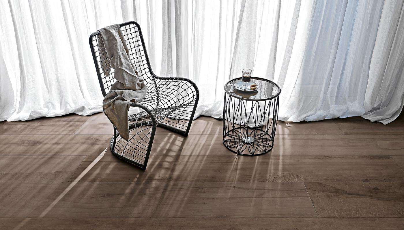 Pokój z podłogą wyłożoną ciemnymi płytkami drewnopodobnymi, z drucianym fotelem, okrągłym stolikiem i białą zasłoną