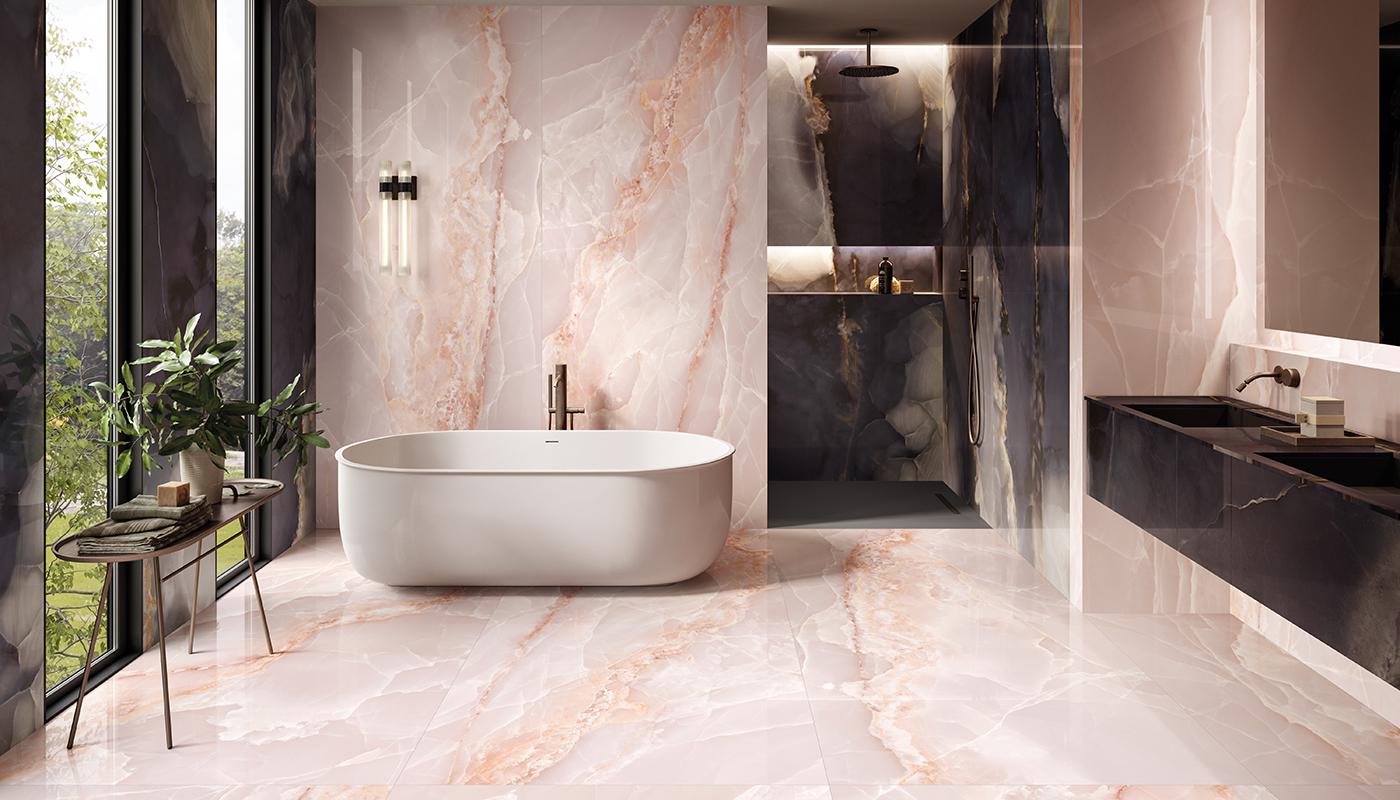 Elegancka łazienka wyłożona różowymi płytkami imitującymi kamień onyks, z białą wanną wolnostojącą, kabiną prysznicową, wiszącą półką z dwiema umywalkami i stoliczkiem z ręcznikiem i kwiatem