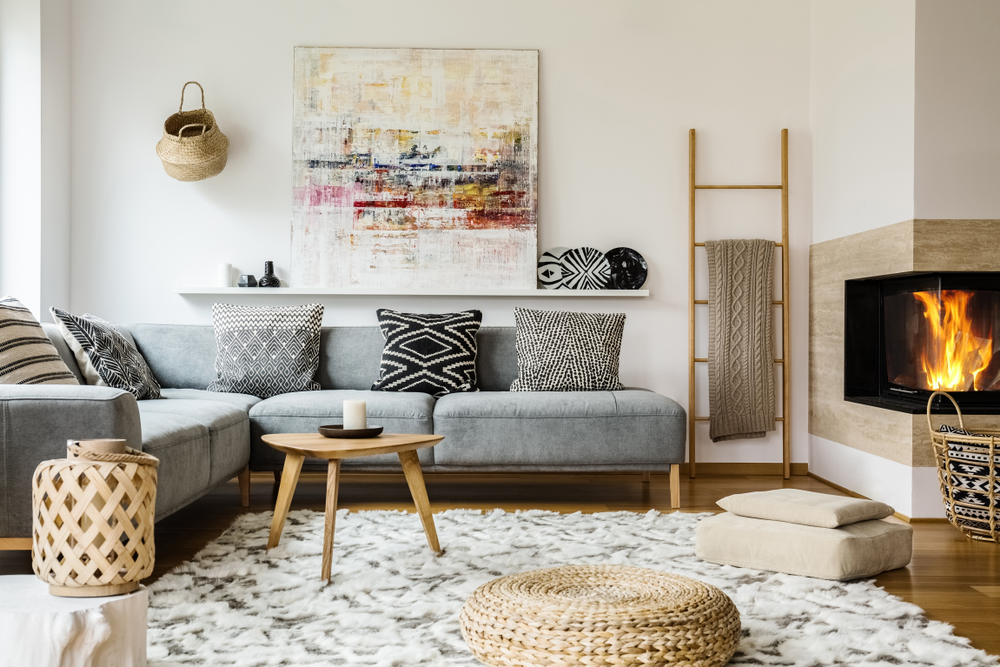Przytulny salon w stylu skandynawskim z szarą kanapą-narożnikiem z poduszkami, puszystym dywanem, kominkiem, trójkątnym, drewnianym stoliczkiem, obrazem, ozdobami i wiklinowymi dodatkami