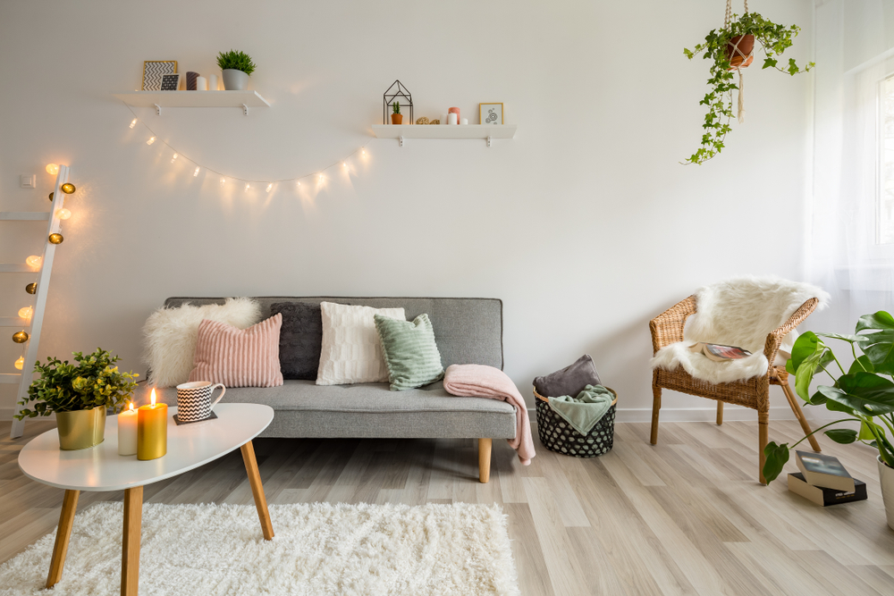 Przytulny salon w stylu skandynawskim z szarą kanapą z kolorowymi poduszkami, fotelem wiklinowym, trójkątnym stoliczkiem na białym, puszystym dywanie, z lampkami, kwiatami i ozdobami