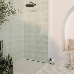 Jasna łazienka ze ścianą pod natryskiem wyłożoną jasnozielonymi cegiełkami w połysku Rebels Sage Gloss z deszczownicą, kosmetykami na podłodze i dużymi roślinami