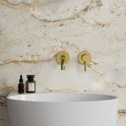 Widok od przodu na białą umywalkę ze złotą baterią podtynkową Omnires Y na tle jasnej, beżowej ściany z kamiennym wzorem