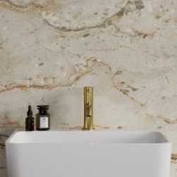 Widok od przodu na białą umywalkę ze złotą baterią stojącą Omnires Y na tle jasnej, beżowej ściany z kamiennym wzorem