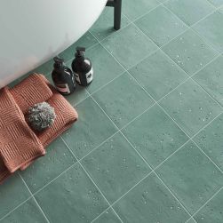 Widok na podłogę w łazience wyłożoną zielonymi płytkami dekoracyjnymi Stardust Pebbles Teal, z białą wanną, ręcznikiem i kosmetykami