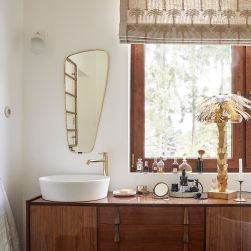Romantyczna łazienka z drewnianą szafką vintage z białą umywalką nablatową i baterią stojącą złotą Omnires Y, z dużą ilością kosmetyków, ciekawą lampką, lustrem oraz oknem z roletą