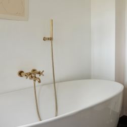 Złota bateria wannowa ścienna ze słuchawką prysznicową z kolekcji Omnires Modern przymocowana do białej ściany nad białą wanną owalną