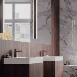 Fragment łazienki z jedną ścianą ciemnobrązową drewnianą, a drugą jasną marmurową, z dwiema szafkami drewnianymi z białymi umywalkami i bateriami stojącymi w chromie Omnires Fresh oraz dużym lustrem