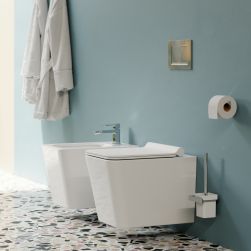 Pastelowa łazienka z kolorowymi płytkami lastryko na podłodze, białą miską WC wiszącą i bidetem z baterią w chromie Slide, z uchwytem na papier toaletowy i szczotką toaletową oraz białym szlafrokiem zawieszonym na ścianie