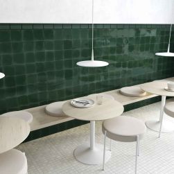 Restauracja z białą podłogą i ciemnozielonymi cegiełkami w połysku Fayenza Belt Royal Green Gloss na ścianie, z drewnianą ławą, okrągłymi stolikami, taboretami i lampami wiszącymi