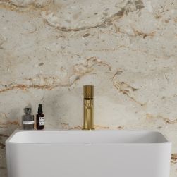 Widok od przodu na białą umywalkę ze złotą baterią stojącą Omnires Contour na tle beżowej, kamiennej ściany