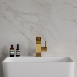 Widok od przodu na białą umywalkę ze złotą baterią stojącą Omnires Darling na tle jasnej, marmurowej ściany