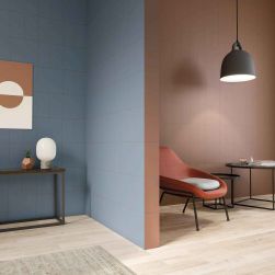 Pomieszczenie podzielone na dwie części ze ścianą w drugiej wyłożoną brązowymi płytkami bazowymi Solid L Cotto, z pomarańczowym fotelem, okrągłymi stolikami i lampą wiszącą