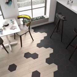 Widok na podłogę w kuchni wyłożoną drewnopodobnymi płytkami w kształcie jodełki Chevron B Wood Light z czarnymi płytkami heksagon, z białym stolikiem z krzesłami i wyspą z czarnymi taboretami