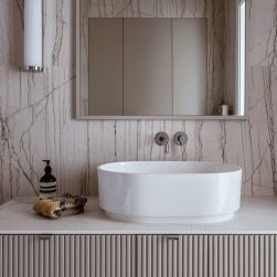 Fragment romantycznej łazienki z marmurową ścianą, pastelową szafką z białą, owalną umywalką nablatową Omnires Cadence, baterią podtynkową, lustrem i kinkietem