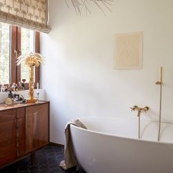 Rustykalna łazienka z drewnianą szafką pod oknem z roletą z ozdobami i kosmetykami, z białą wanną owalną ze złotą baterią wannową ze słuchawką prysznicową z kolekcji Omnires Modern oraz z obrazkiem na ścianie