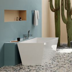Kolorowa łazienka z podłogą lastryko, niebieską i żółtą ścianą, dużymi kaktusami, białą wanną prostokątną przy półce z zamontowaną baterią nawannową w chromie z kolekcji Omnires Slide i białym ręcznikiem wiszącym na ścianie