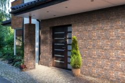 Przytulny dom w otoczeniu zieleni, z brązowymi drzwiami wejściowymi i kamieniem elewacyjnym Lagos Copper