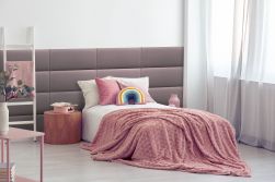 Dziecięca sypialnia z łóżkiem z różową narzutą i pastelowymi poduszkami, ścianą wyłożoną w części różowymi panelami tapicerowanymi Softi, oknem ze zwiewną zasłoną, okrągłym stolikiem, drabinką i różowym stolikiem
