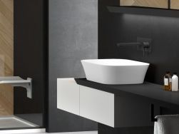Fragment łazienki utrzymanej w szarej i czarnej kolorystyce z półką wiszącą z białą szafką i białą umywalką nablatową oraz czarną baterią podtynkową Besco Decco, lustrem i kabiną prysznicową