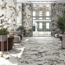 Hotelowa restauracja wyłożona płytkami imitującymi marmur z kolekcji Les Bijoux ze stolikami, wygodnymi fotelami, dużymi kwiatami w donicach, lampami wiszącymi i szklanymi drzwiami