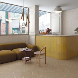 Bar z podłogą wyłożoną brązowymi płytkami lastryko Croccante-R Nuez z żółtą kanapą, okrągłym stolikiem, krzesłem i lampą wiszącą
