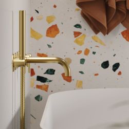 Widok z boku na baterię wannową podtynkową 5-otworową z kolekcji Y w kolorze złota szczotkowanego na tle ściany wyłożonej kolorowymi płytkami lastryko i nad białą wanną