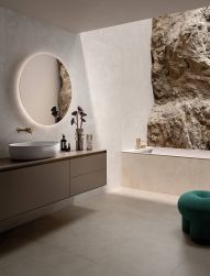 Łazienka ze ścianą wyłożoną jasnymi płytkami imitującymi beton All Around Concrete White, z zabudowaną wanną, szafką wiszącą z umywalką nablatową i okrągłym lustrem
