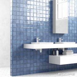 Łazienka z białą podłogą i ścianą na środku wyłożoną niebieskimi cegiełkami z kolekcji Altea, z dwiema białymi półkami wiszącymi, z dwiema umywalkami z bateriami stojącymi i lustrem z szafką