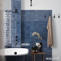 Łazienka ze ścianą wyłożoną niebieskimi cegiełkami z kolekcji Altea z biało-czarną wanną, baterią podtynkową i deszczownicą, taboretem z kosmetykami oraz ręcznikiem na wieszaku