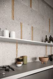 Ściana w kuchni wyłożona szarymi płytkami imitującymi kamień z kolekcji Ego Grigio z drewnianymi wstawkami, z szarymi meblami, wiszącą półką i naczyniami