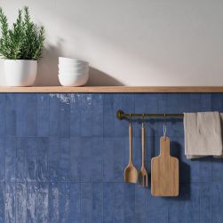 Fragment kuchni ze ścianką z półką wyłożoną niebieskimi cegiełkami w połysku Aqua Blue z drewnianym naczyniami na wieszaku, białymi miskami i kwiatem w donicy