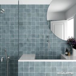 Widok na fragment łazienki z kabiną prysznicową, zabudowaną umywalką z okrągłym lustrem, kwiatami i kosmetykami oraz błękitnymi cegiełkami z kolekcji Argile