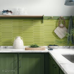 Kolorowa kuchnia ze ścianą wyłożoną jasnymi, zielonymi płytkami z kolekcji Arrow Apple w kształcie strzały, z ciemnymi zielonymi meblami i białymi blatami oraz wiszącą półką