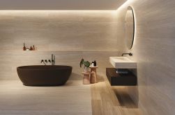 Ciepła łazienka wyłożona beżowymi płytkami imitującymi kamień Arte Desert z brązową wanną owalną, wiszącą umywalką nad brązową półką, okrągłym lustrem, dwoma taboretami i kosmetykami