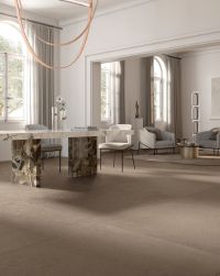 Elegancki i przestronny salon z brązowymi płytkami imitującymi beton Balance Mud na podłodze, z marmurowym stołem, dwoma białymi krzesłami, dwoma fotelami i małym stolikiem