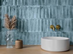 Zbliżenie na ścianę w łazience wyłożoną niebieskimi cegiełkami w połysku Bari Blue z drewnianym blatem, okrągłą umywalką nablatową, baterią podtynkową i szklanym wazonem z dekoracyjnymi liśćmi