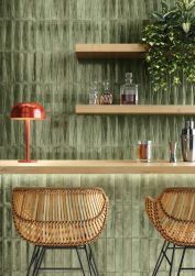 Ściana wyłożona zielonymi cegiełkami w połysku Bari Green z drewnianym blatem i półkami, dwoma wiklinowymi krzesłami, czerwoną lampką, kwiatem i naczyniami z alkoholem