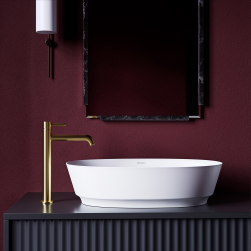 Biała umywalka nablatowa owalna Omnires Neo ze złotą baterią stojącą na czarnej półce, z lustrem w ramie i kinkietem na tle burgundowej ściany