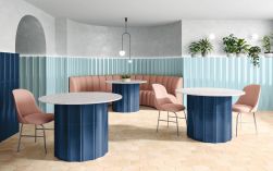 Restauracja ze ścianami wyłożonymi do połowy błękitnymi płytkami trójwymiarowymi Bow Sky z okrągłymi stołami, pudrowymi krzesłami i kanapą, lampą wiszącą i roślinami