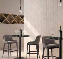 Restauracja z szarymi, okrągłymi stolikami z butelkami wina i kieliszkami, szarymi, wysokimi fotelami, wiszącymi lampami typu żar&oacute;wka, wnęką w ścianie z butelkami wina i płytkami z kolekcji Gravel