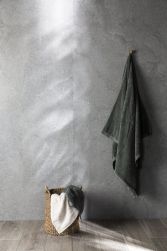 Widok na ścianę wyłożoną szarymi płytkami imitującymi kamień Lucca Grey z ciemnym, zielonym ręcznikiem oraz wiklinowym koszem