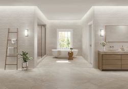 Duża i przestronna łazienka wyłożona beżowymi płytkami imitującymi kamień Lucca Beige z białą wanną owalną pod oknem, drewnianą szafką i lustrem, drabinką z ręcznikami i kwiatem doniczkowym