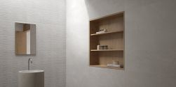 Widok na ściany w łazience wyłożone białymi płytkami imitującymi kamień Lucca Grey z drewnianymi półkami w ścianie z kosmetykami, okrągłą umywalką stojącą oraz lustrem