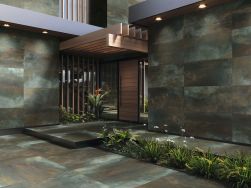 Elewacja domu i chodnik wyłożone płytkami imitującymi metal Metallic Wild z brązowymi drzwiami wejściowymi, zadaszeniem i pasem zieleni