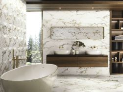 Elegancka łazienka z białą, owalną wanną i baterią wolnostojącą złotą, półką wiszącą drewnianą z dwoma umywalkami nablatowymi, długim lustrem, szafkami z akcesoriami, wysokim oknem oraz płytkami podłogowymi i ściennymi imitującymi marmur z kolekcji Bellag
